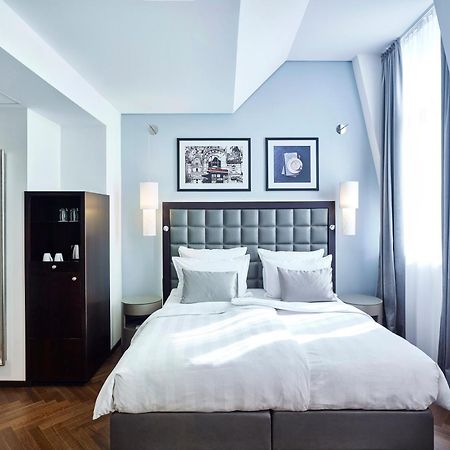 Steigenberger Hotel Herrenhof Відень Екстер'єр фото The photo shows a stylish hotel room with a modern design. It features a large, neatly made bed with white linens and gray accents. On the wall above the bed, there are two framed artworks. The room has large windows with light gray curtains, allowin