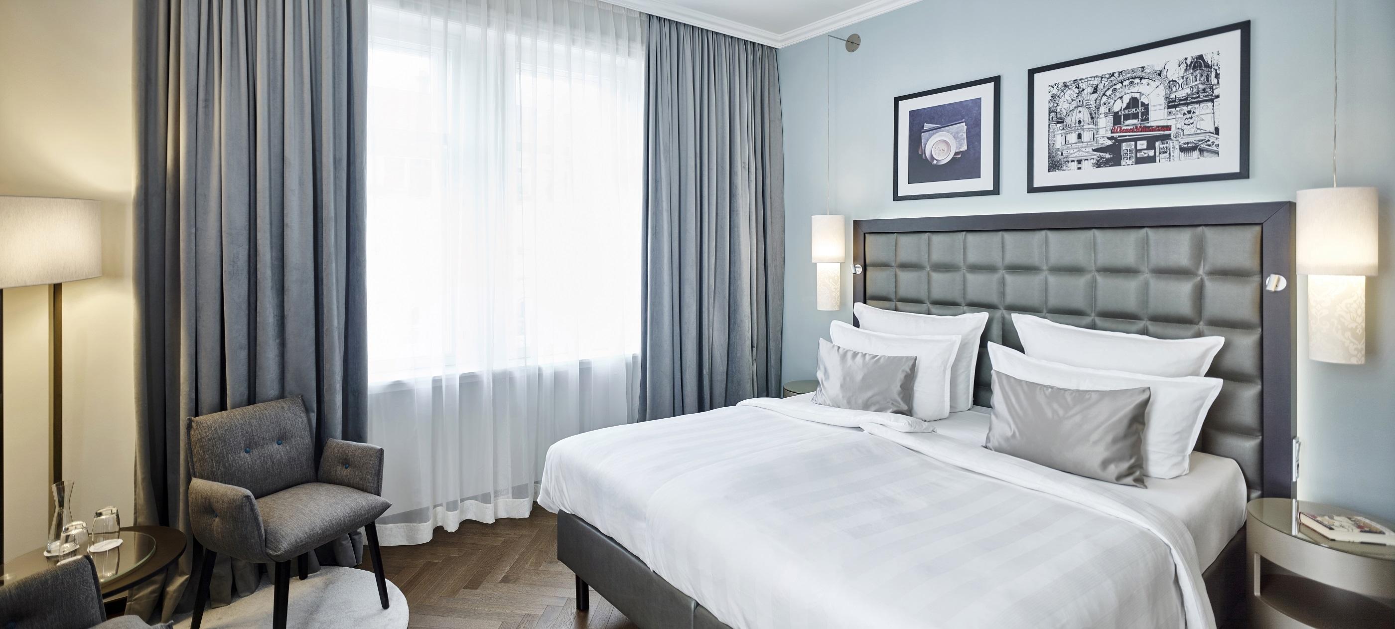 Steigenberger Hotel Herrenhof Відень Екстер'єр фото The photo shows a modern hotel room featuring a neatly made double bed with white bedding and decorative gray pillows. Behind the bed, there is a stylish, tufted headboard. To one side, there's a comfortable armchair next to a small round table. The 