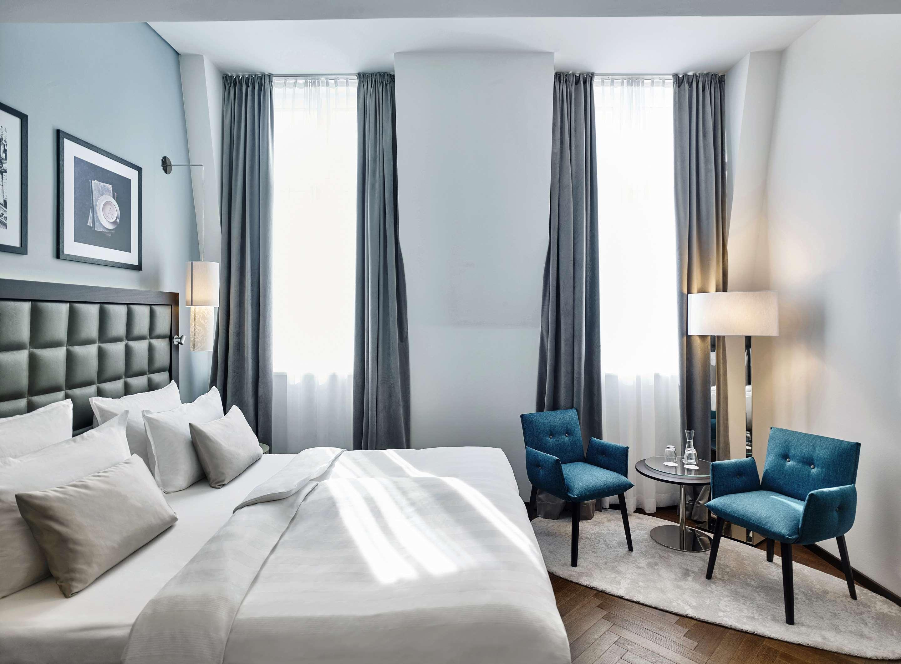 Steigenberger Hotel Herrenhof Відень Екстер'єр фото The photo shows a stylish hotel room featuring a neatly made bed with multiple pillows and a light-colored bedspread. Behind the bed, there is a dark, tufted headboard. Large windows dressed with long, gray curtains allow natural light to brighten th