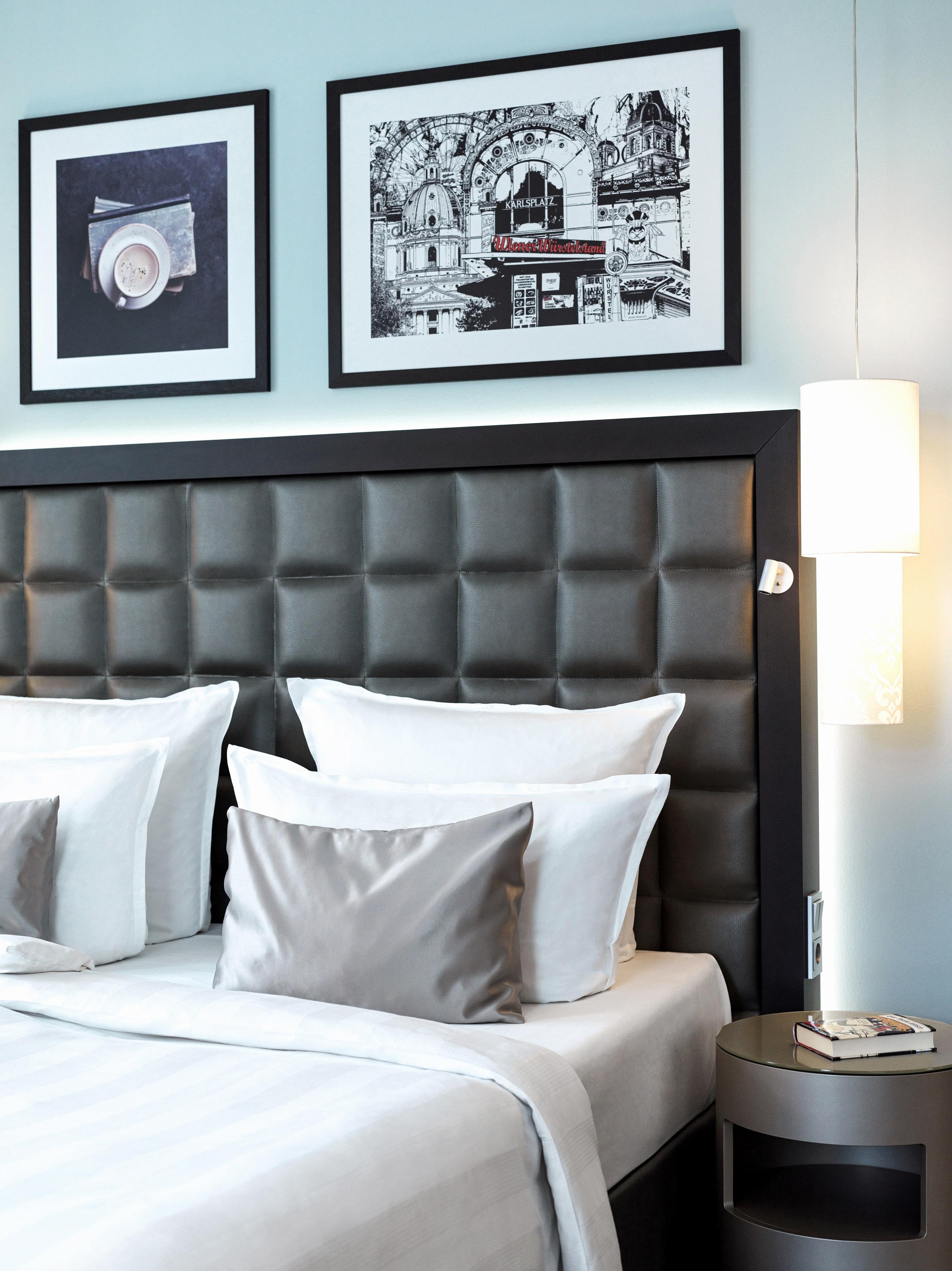 Steigenberger Hotel Herrenhof Відень Екстер'єр фото The image displays a modern hotel room setup. It features a bed with a stylish headboard that has a tufted design in dark leather or faux leather. The bed is adorned with several white and silver decorative pillows. Above the bed, there are framed bl