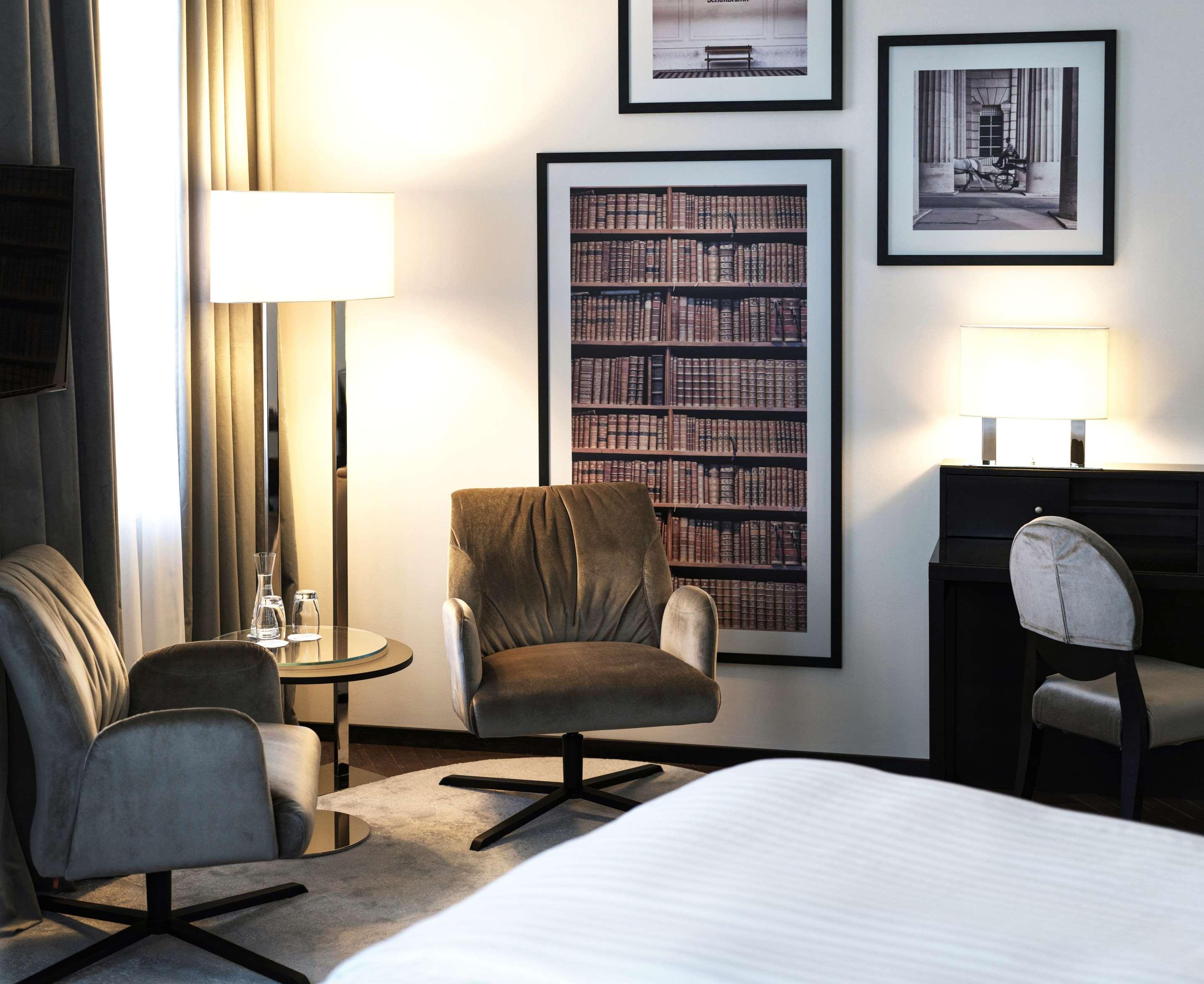Steigenberger Hotel Herrenhof Відень Екстер'єр фото The photo shows a modern hotel room or lounge area. It features two stylish chairs, a small round glass table, and a standing lamp adding warm light to the space. Behind the chairs, there is a large framed print that resembles a bookshelf filled with