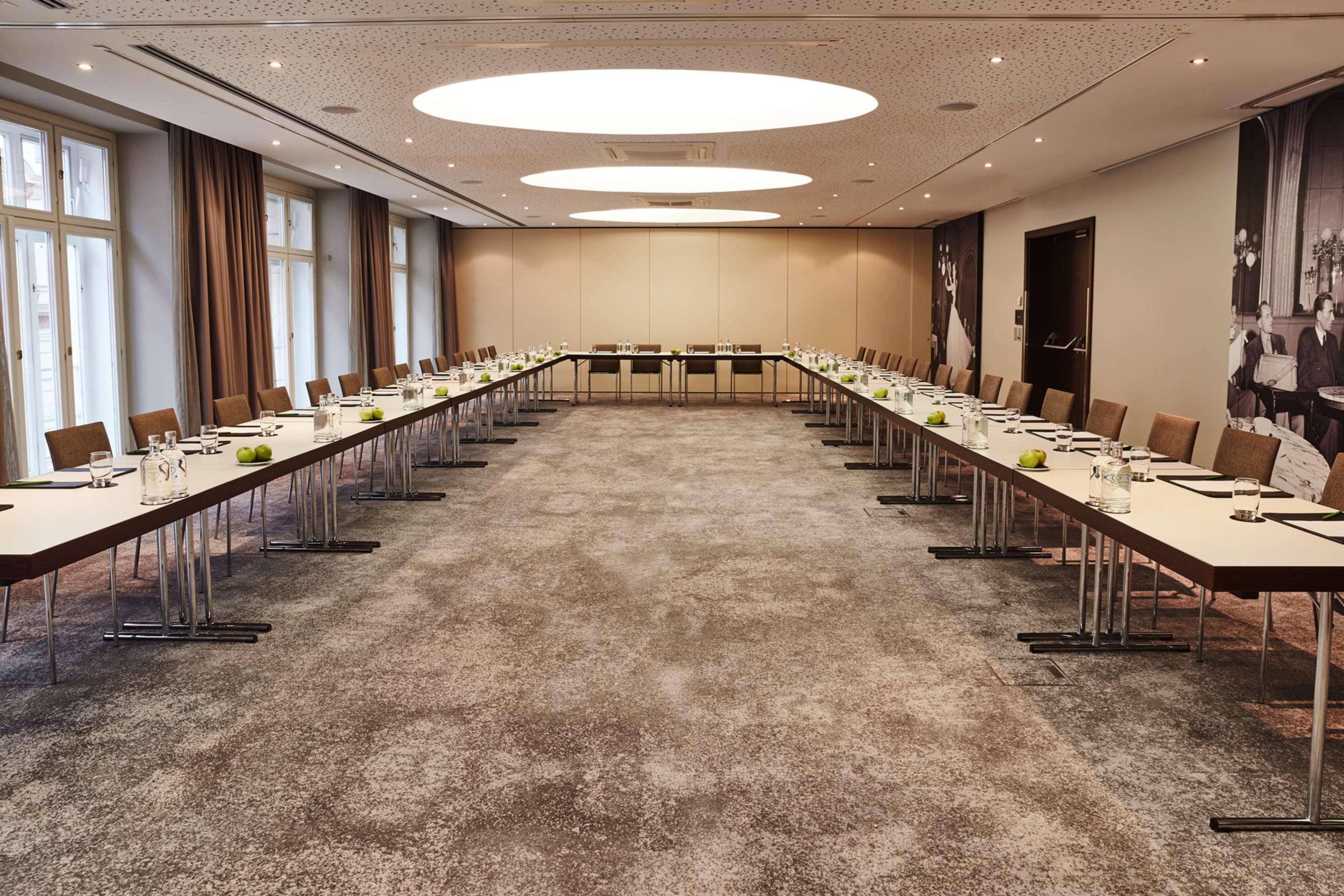 Steigenberger Hotel Herrenhof Відень Екстер'єр фото The photo shows a spacious conference room set up for a meeting. There are two long tables arranged in a U-shape, each covered with white tablecloths. The tables are equipped with glasses, small notepads, and pens for attendees. The room features lar