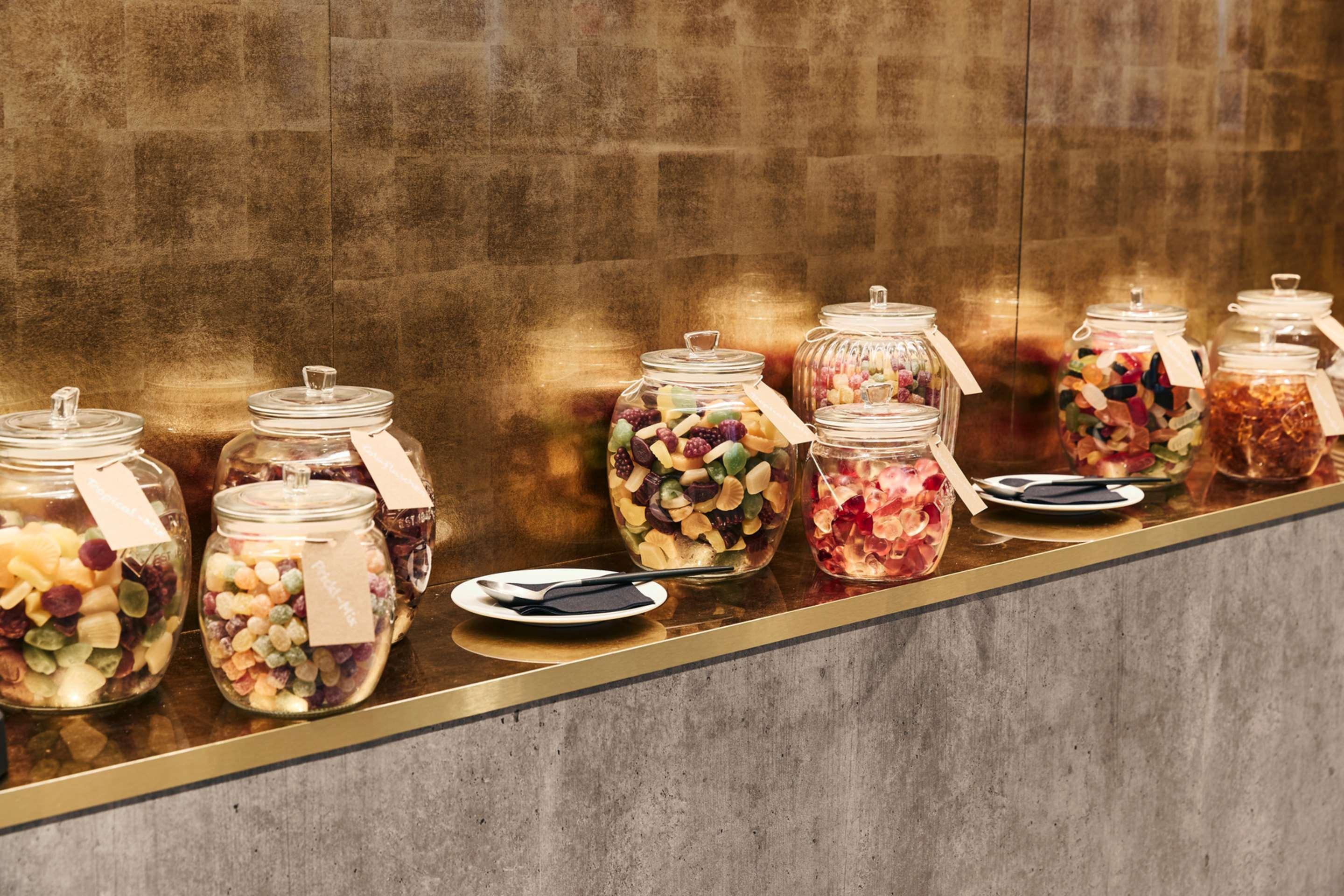 Steigenberger Hotel Herrenhof Відень Екстер'єр фото The photo shows a display of several glass jars filled with various types of candy. Each jar is labeled and positioned on a sleek, horizontal surface against a golden background. There are some small plates placed in front of the jars, which likely c