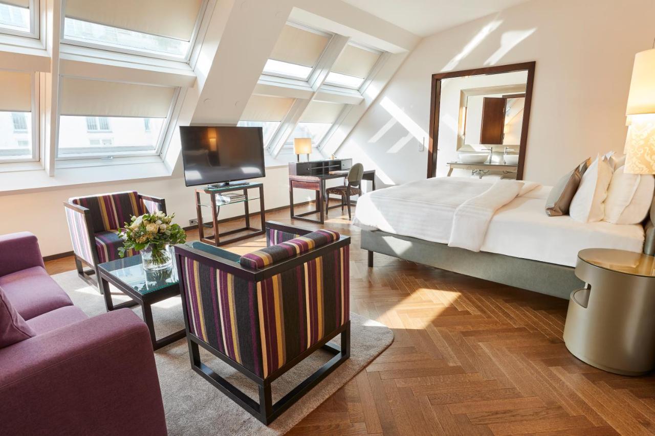 Steigenberger Hotel Herrenhof Відень Екстер'єр фото The photo shows a hotel room with a modern and stylish design. It features a large bed with white bedding, situated against a wall. In front of the bed, there's a contemporary coffee table with a small floral arrangement. Two chairs with a colorful s