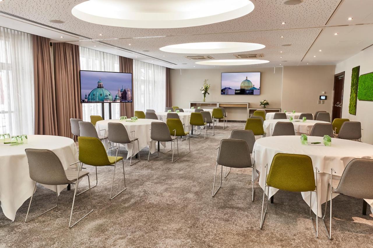 Steigenberger Hotel Herrenhof Відень Екстер'єр фото The photo depicts a modern conference or meeting room. There are several round tables covered with white tablecloths, each paired with chairs—some in shades of gray and others with green accents. The room is well-lit with large windows allowing natur