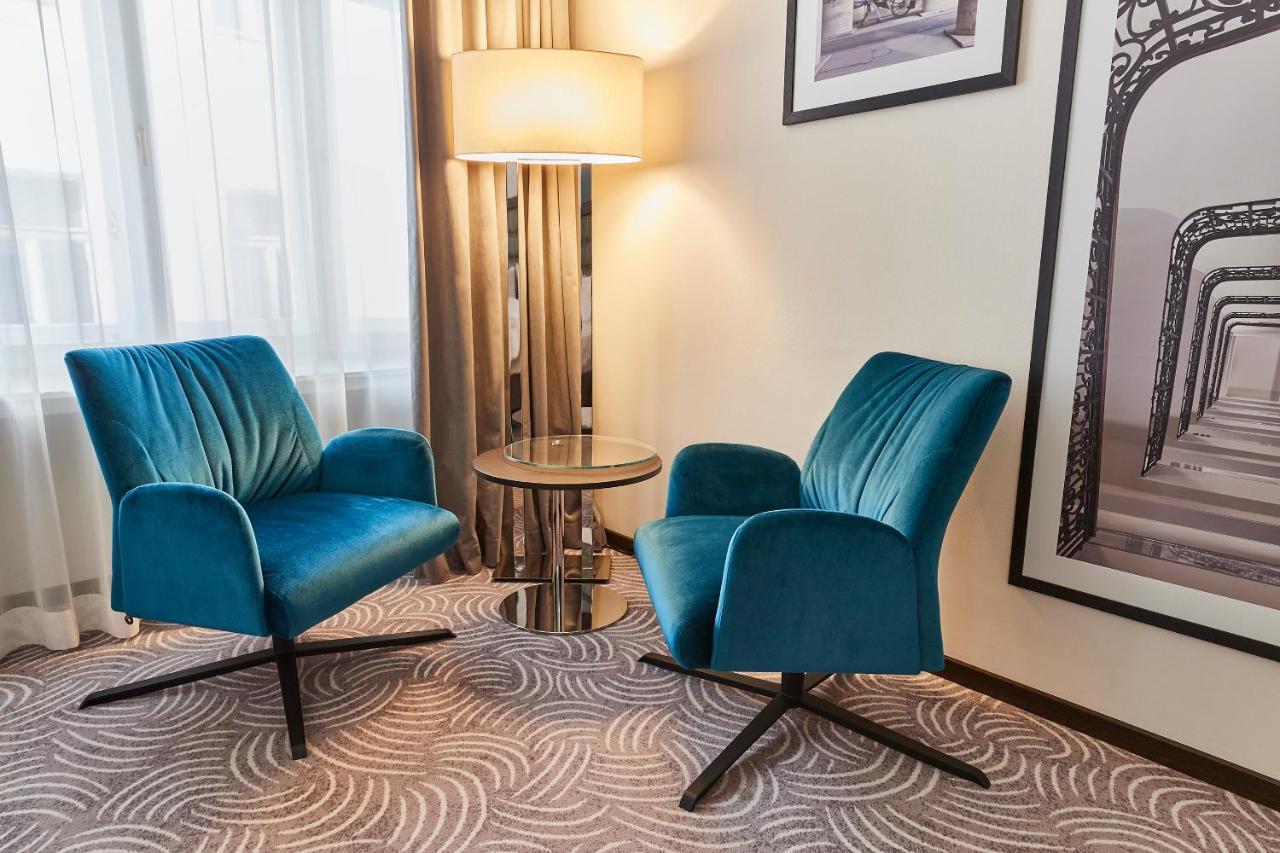Steigenberger Hotel Herrenhof Відень Екстер'єр фото The photo shows a cozy corner of a room featuring two modern, teal upholstered chairs positioned around a small round table. The chairs have a stylish design with a curved back and are set on a black, star-shaped base. Next to the chairs is a floor l