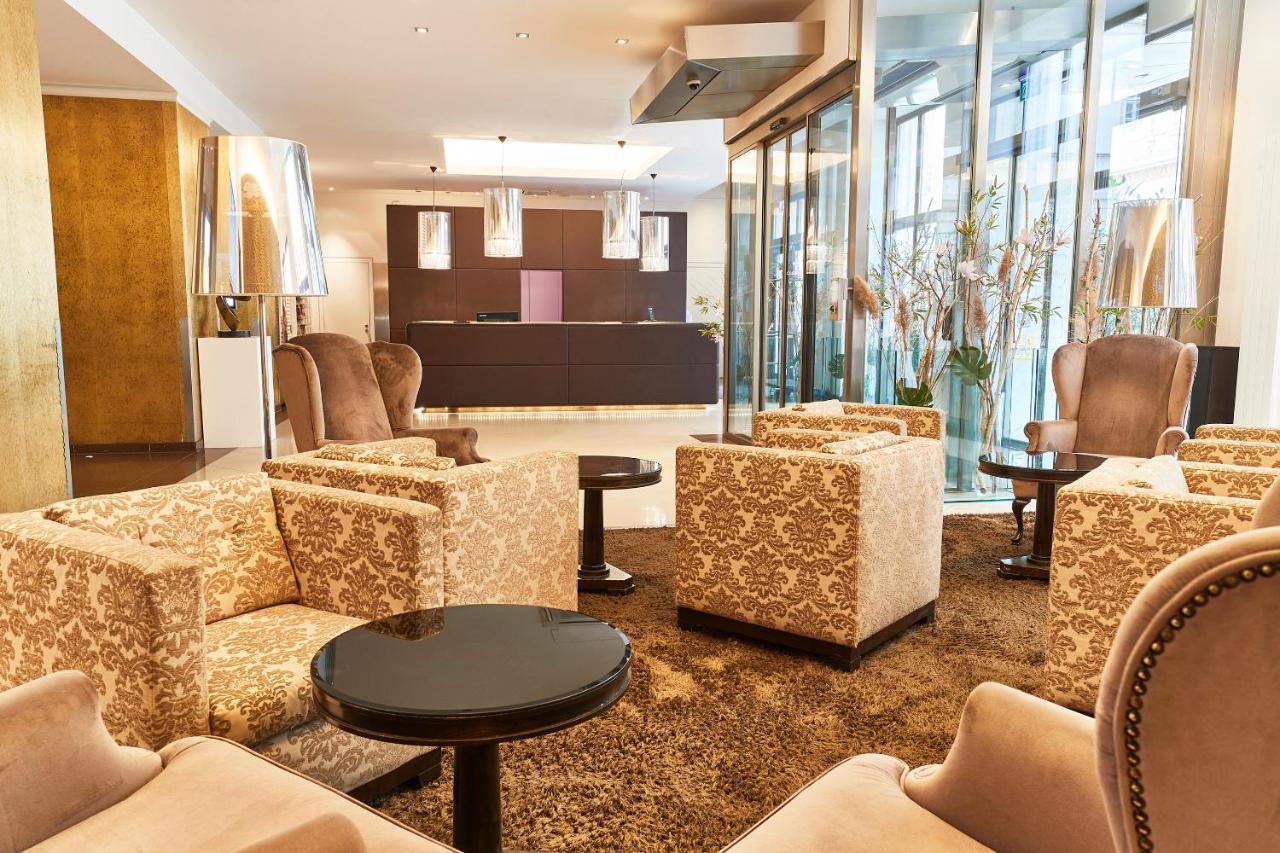 Steigenberger Hotel Herrenhof Відень Екстер'єр фото The photo shows a hotel lobby or waiting area designed with a modern and elegant style. There are several plush, patterned armchairs arranged around small round tables. The flooring is covered with a large, soft rug. In the background, there is a rec