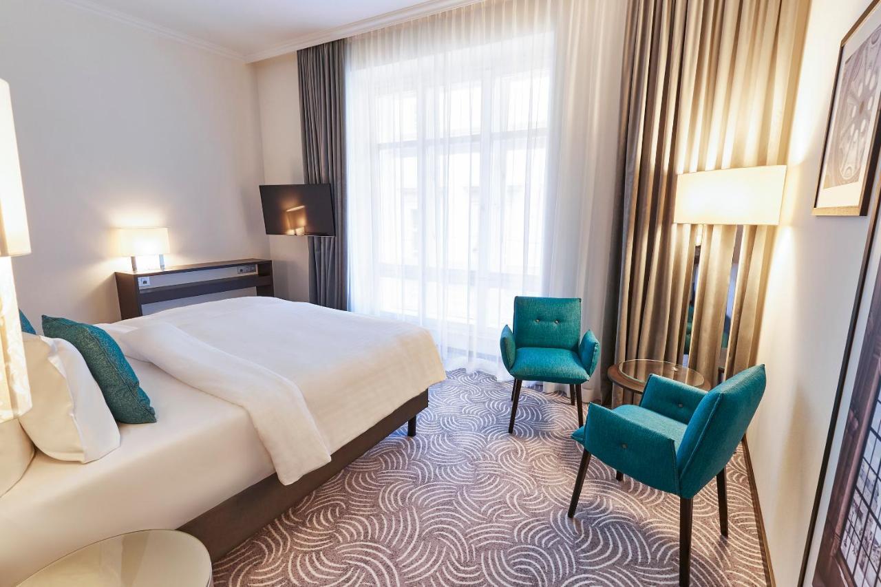 Steigenberger Hotel Herrenhof Відень Екстер'єр фото The photo shows a modern hotel room designed with light colors and contemporary furnishings. There is a large bed with white bedding, accented by teal decorative pillows. To one side, there are two teal chairs and a small glass table, creating a cozy