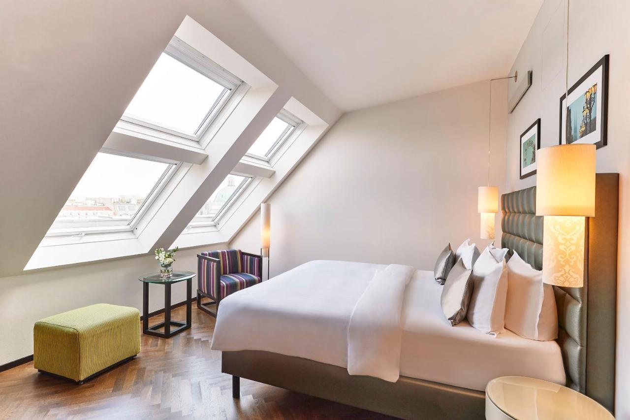 Steigenberger Hotel Herrenhof Відень Екстер'єр фото The photo shows a modern and stylish hotel room. It features a comfortable bed with neatly arranged pillows, a small seating area with a chair, and a side table next to it. The room has large slanted windows that allow natural light to fill the space