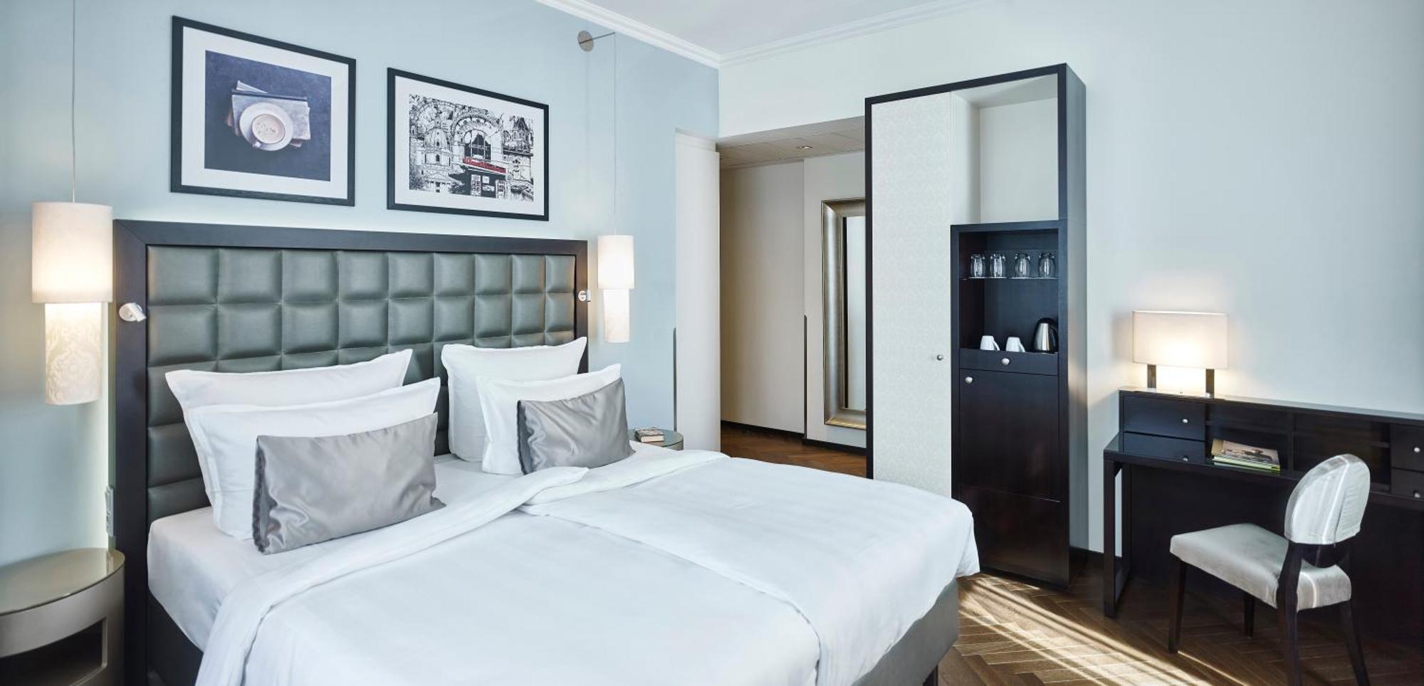 Steigenberger Hotel Herrenhof Відень Екстер'єр фото The photo shows a modern hotel room featuring a double bed with white linens and gray accent pillows. The headboard is upholstered in a tufted design. On the wall behind the bed, there are framed artworks, adding a decorative touch. To the right, the