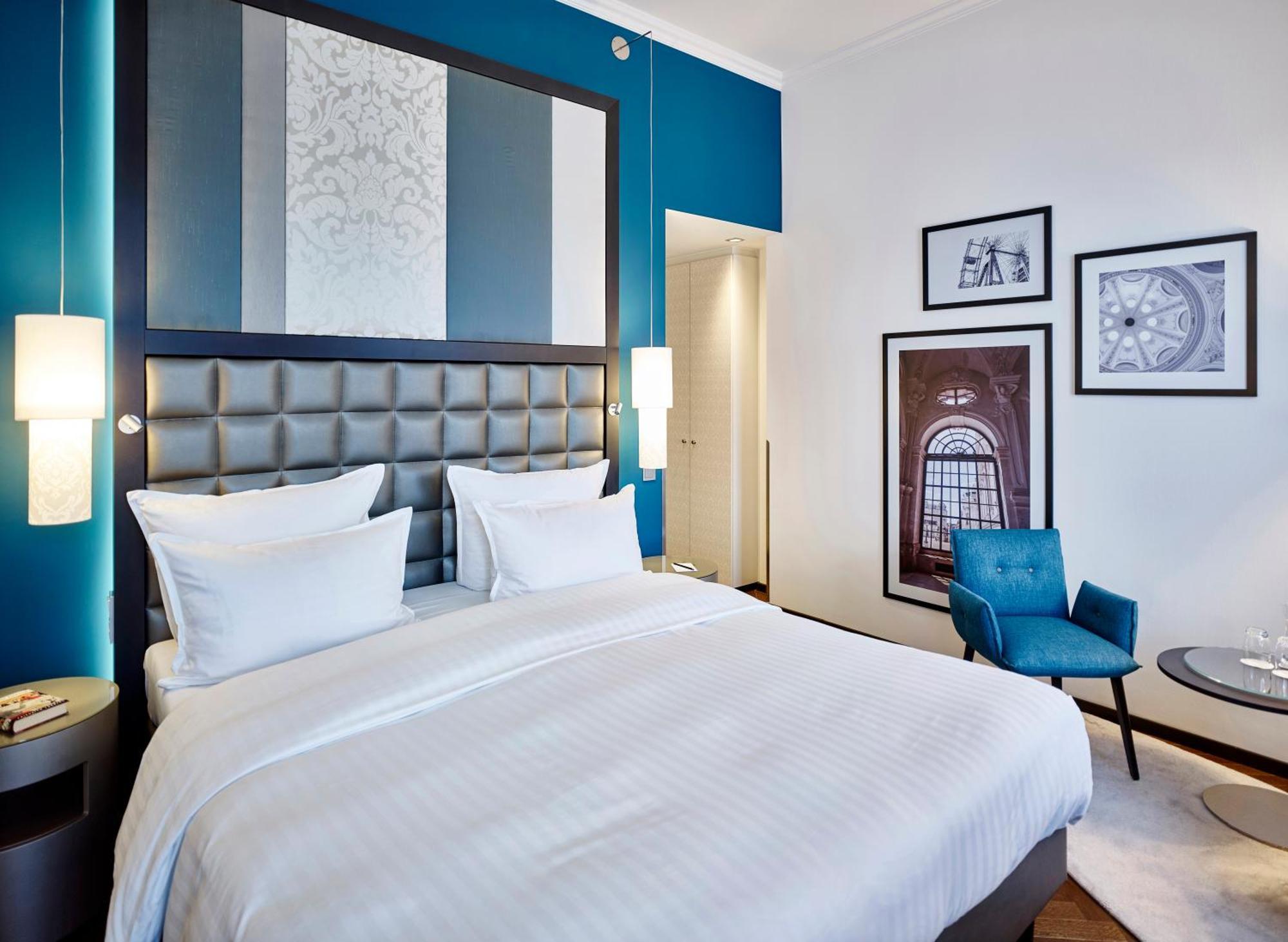 Steigenberger Hotel Herrenhof Відень Екстер'єр фото The photo shows a modern hotel room featuring a neatly made bed with white linens and two pillows. The wall behind the bed has a decorative design with alternating colors of blue and white. On either side of the bed, there are sleek bedside lamps. To