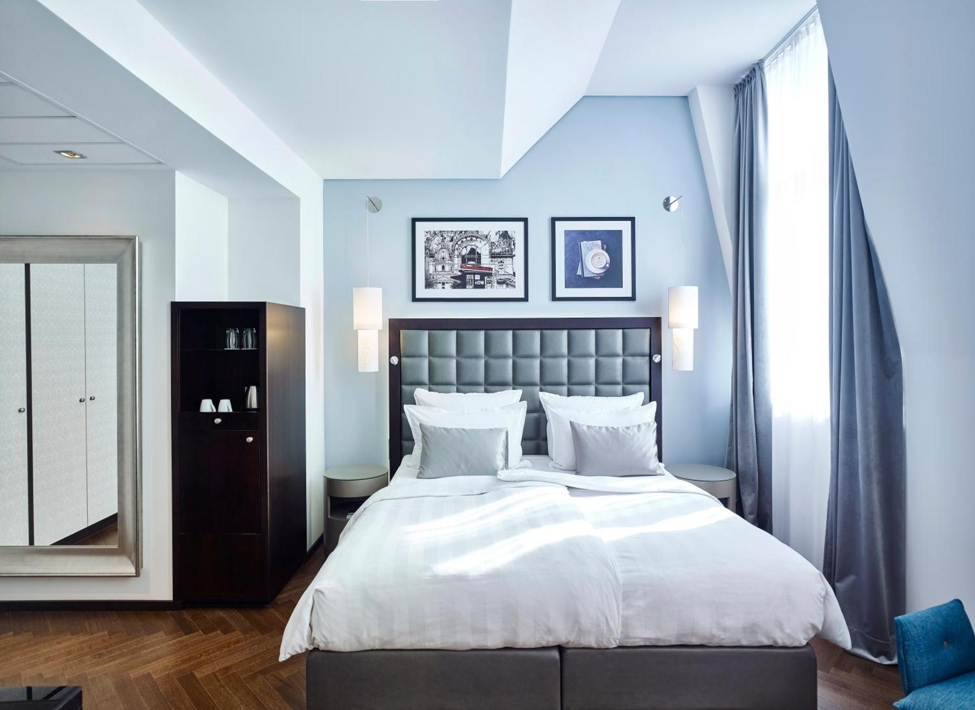Steigenberger Hotel Herrenhof Відень Екстер'єр фото The photo shows a stylish hotel room with a modern design. It features a large, neatly made bed with white linens and gray accents. On the wall above the bed, there are two framed artworks. The room has large windows with light gray curtains, allowin