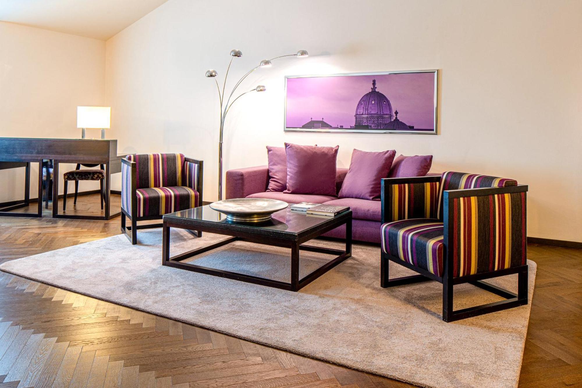 Steigenberger Hotel Herrenhof Відень Екстер'єр фото The photo shows a modern and stylish living room setup. It features a plush purple sofa with several matching cushions. In front of the sofa is a sleek coffee table with a glass top, resting on a soft area rug. Flanking the sofa are two striped armch