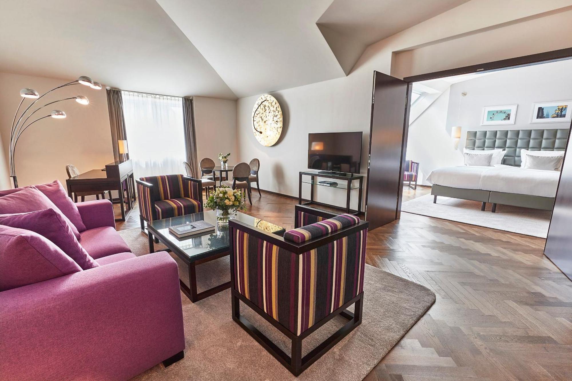 Steigenberger Hotel Herrenhof Відень Екстер'єр фото The photo shows a modern, stylish living space that appears to be part of a hotel suite. The room features a cozy seating area with a purple sofa and two striped chairs around a glass coffee table. A flat-screen TV is mounted on the wall. Large windo