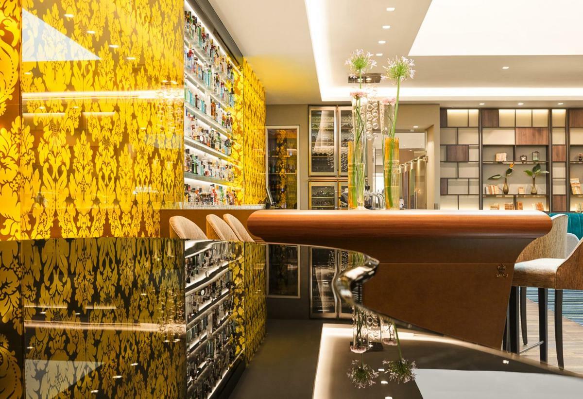 Steigenberger Hotel Herrenhof Відень Екстер'єр фото The photo depicts a modern interior space, likely a bar or lounge. The wall in the background features a striking yellow patterned design, adding a vibrant touch to the environment. There is a sleek, curved bar counter made of wood with stylish bar s